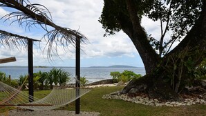 Uma rede sob uma pérgola de madeira com vista para uma praia, costa rochosa e o mar, com ilhas ao longe.