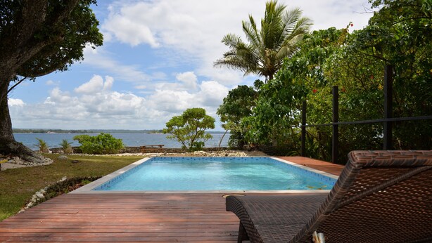 Um deck de madeira com piscina e espreguiçadeiras, com vista para um corpo d'água cercado por árvores e céu claro.