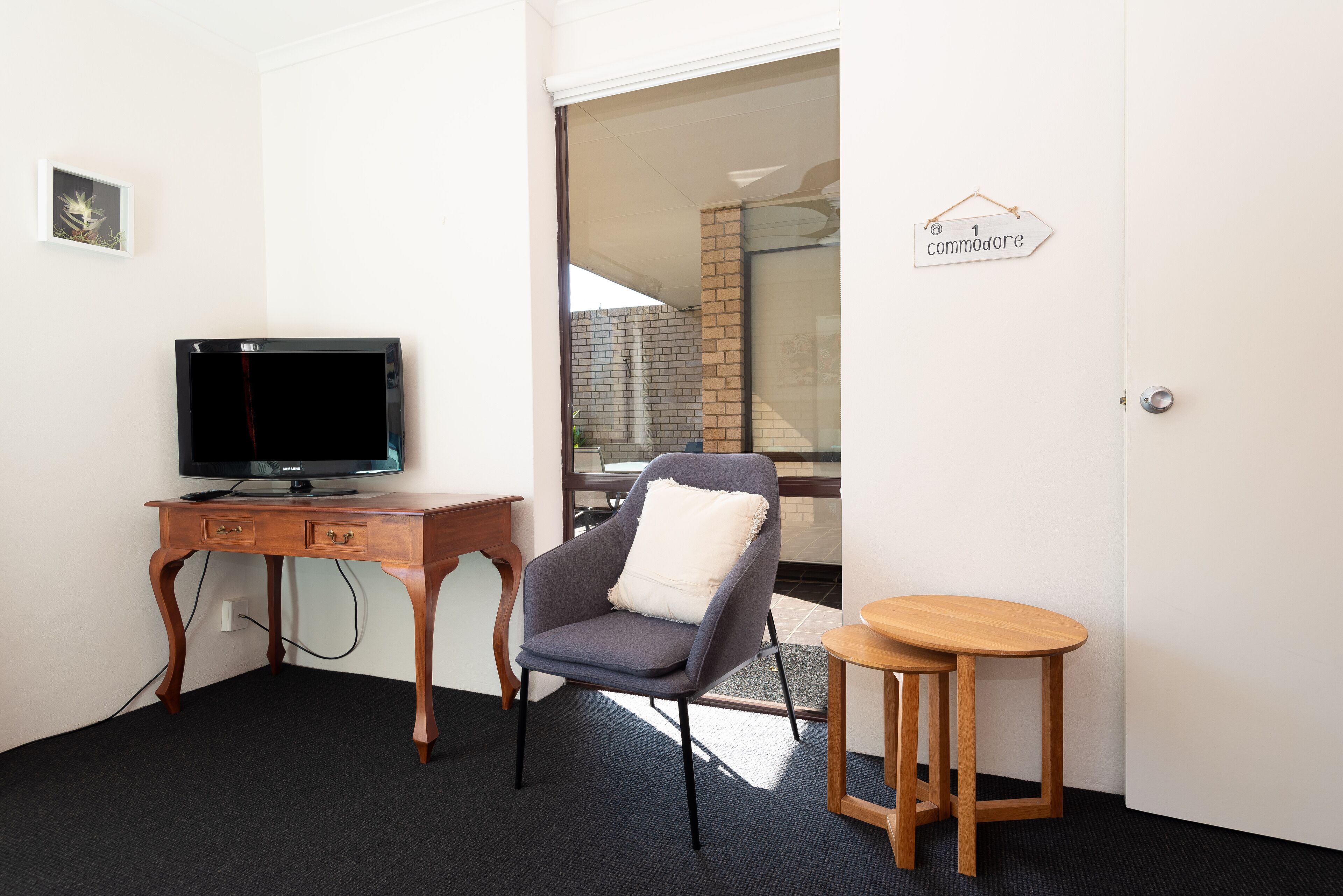Una habitación con un escritorio de madera, una televisión, un sillón gris y un taburete pequeño de madera.