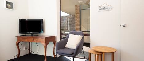 Una habitación con un escritorio de madera, una televisión, un sillón gris y un taburete pequeño de madera.