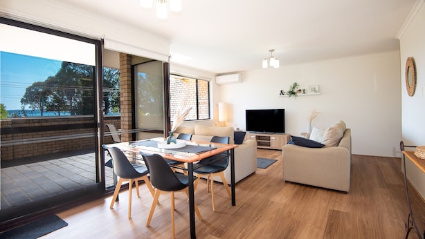 Ett modernt vardagsrum med en matplats, en soffa, en TV och en balkong med utsikt över träd och en byggnad.