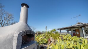 Ein Backofen aus Ziegelsteinen mit Schornstein, umgeben von Pflanzen und einem Haus mit Satellitenschüssel.