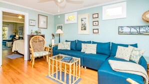 A living room with a blue sofa, a wicker chair, a glass coffee table, and framed pictures on the wall. - Our 'Someday' Cottage - Second Row (Surf City)