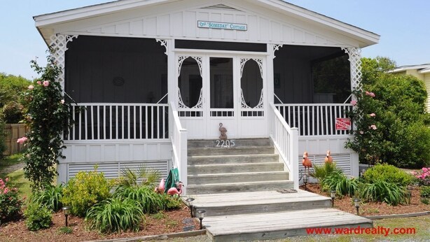 A white cottage with a porch, labeled "One Someday Cottage," featuring a front entrance with steps and a mailbox. - Our 'Someday' Cottage - Second Row (Surf City)