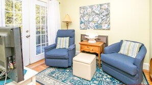 A living room with two blue armchairs, a beige ottoman, a side table with a lamp, and a painting on the wall. - Our 'Someday' Cottage - Second Row (Surf City)