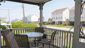 A covered outdoor area with a round table and chairs, a wooden bench, and a view of residential buildings. - Our 'Someday' Cottage - Second Row (Surf City)