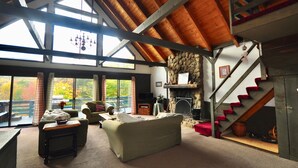 A spacious living room with a high ceiling, large windows, a fireplace, and a staircase.