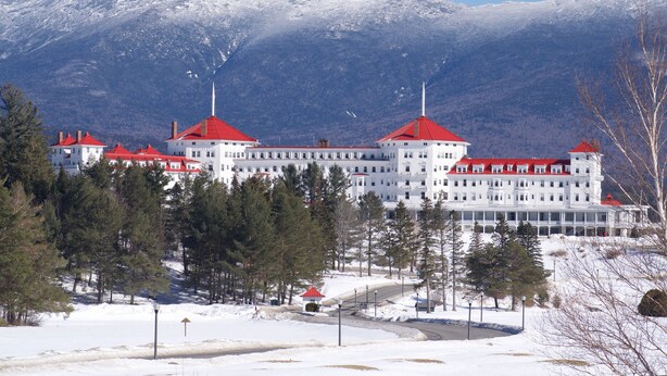 A large, white building with red roofs, surrounded by snow-covered mountains and trees.