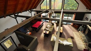 A spacious loft-style room with a high ceiling, a billiards table, a fireplace, and large windows offering a view of the outdoors.