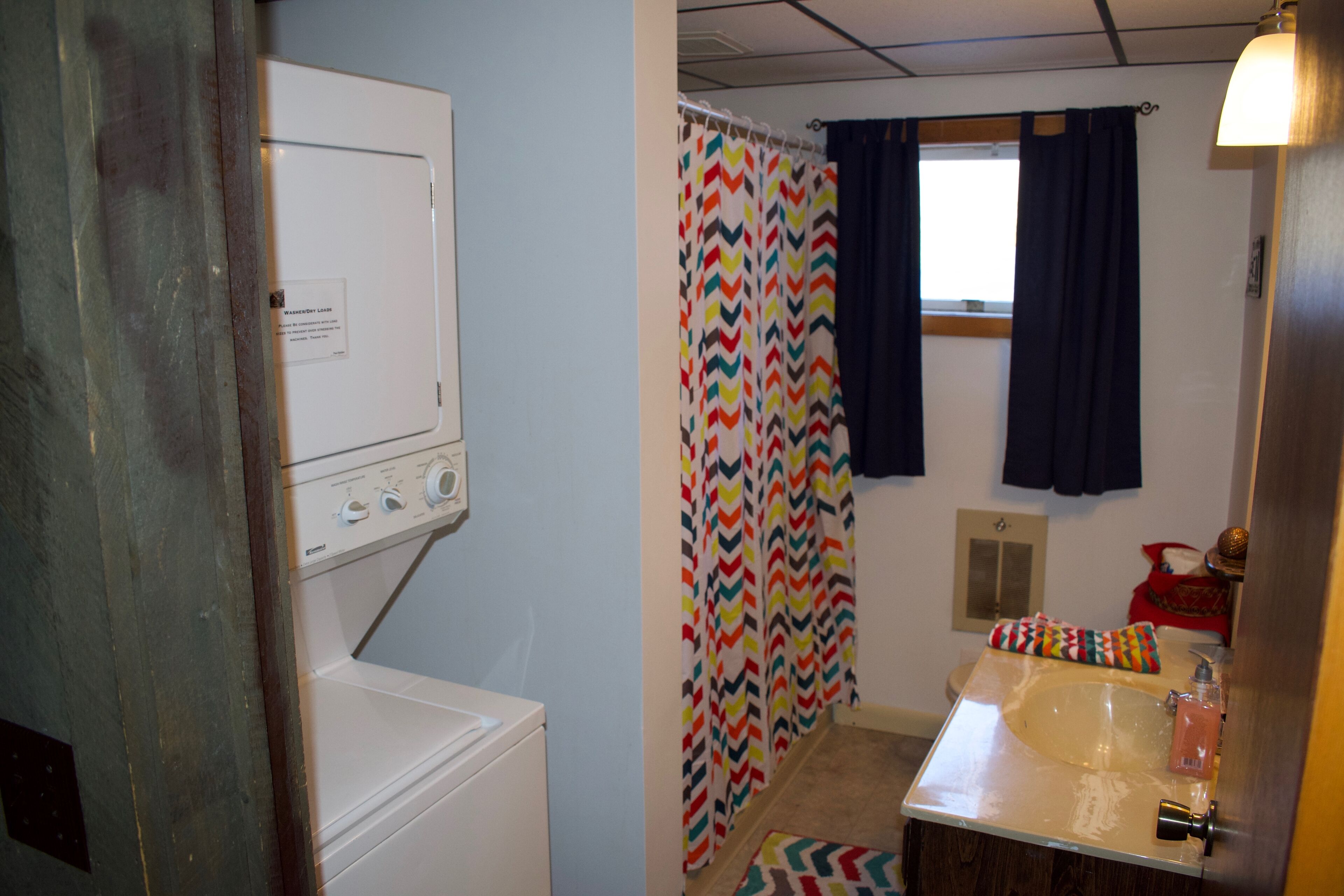 Une petite salle de bain équipée d’un lave-linge, d’un lavabo avec un miroir et d’une douche agrémentée d’un rideau aux motifs colorés en forme de chevrons.