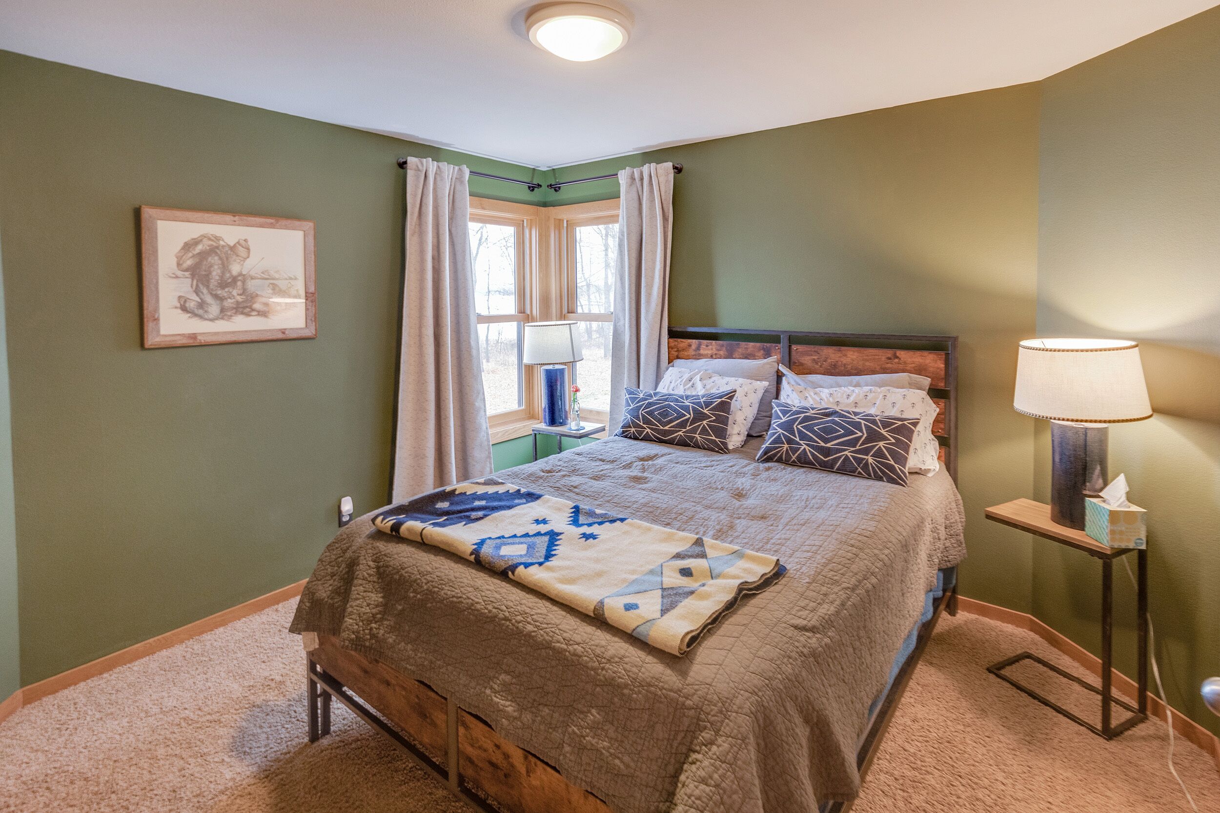 Une chambre à coucher avec un lit, une table de chevet, une lampe, une fenêtre avec des rideaux et un tableau encadré au mur.