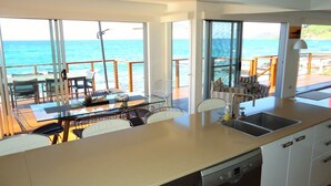 A modern kitchen with large windows, a dining area with a glass table, and a view of the ocean. - Seventeen Steps To The Beach (Boat Harbour Beach)