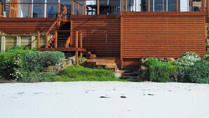 A beachside building with a wooden staircase leading to a deck, surrounded by greenery and a sandy beach. - Seventeen Steps To The Beach (Boat Harbour Beach)