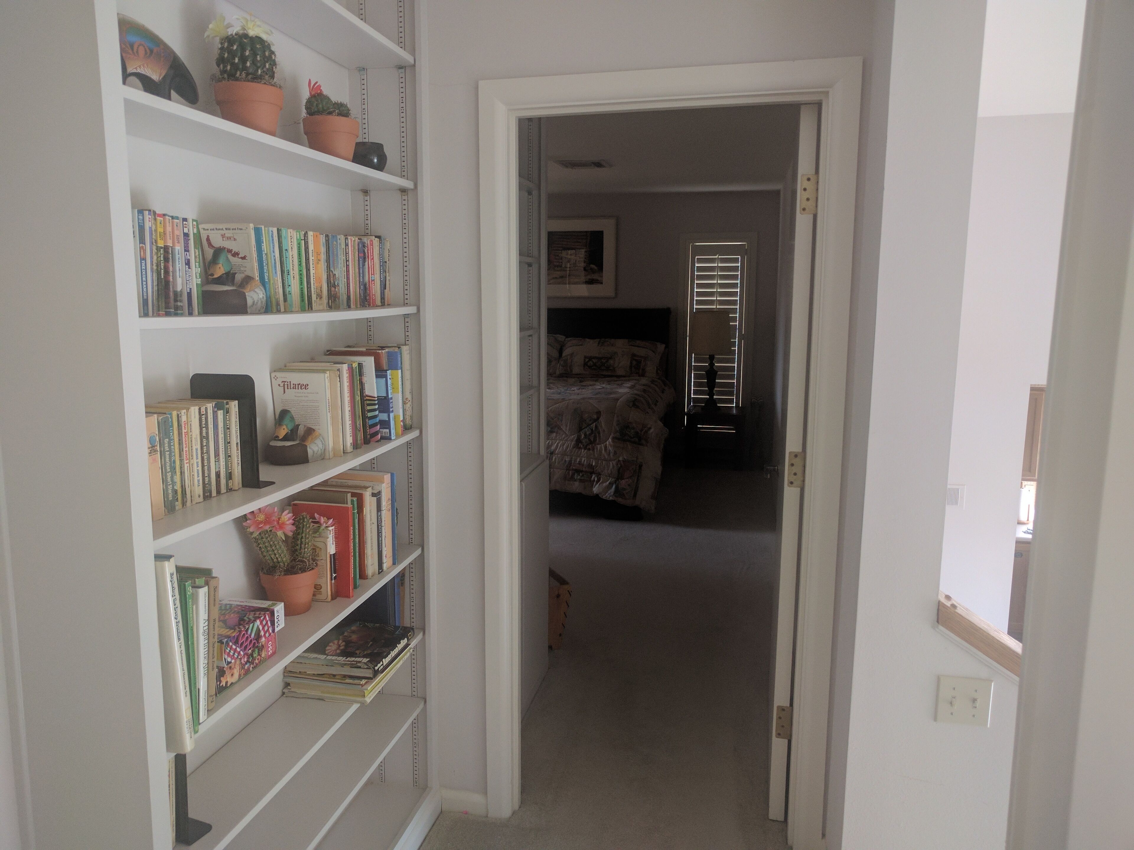 Un couloir mène à une chambre à coucher qui contient un lit, une bibliothèque remplie de livres et d’objets décoratifs, ainsi qu’une fenêtre avec des stores.