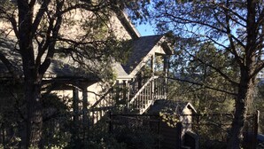 Une grande maison de style rustique, dotée d’un escalier imposant menant à un deuxième étage, entourée d’arbres denses et d’un jardin clos.