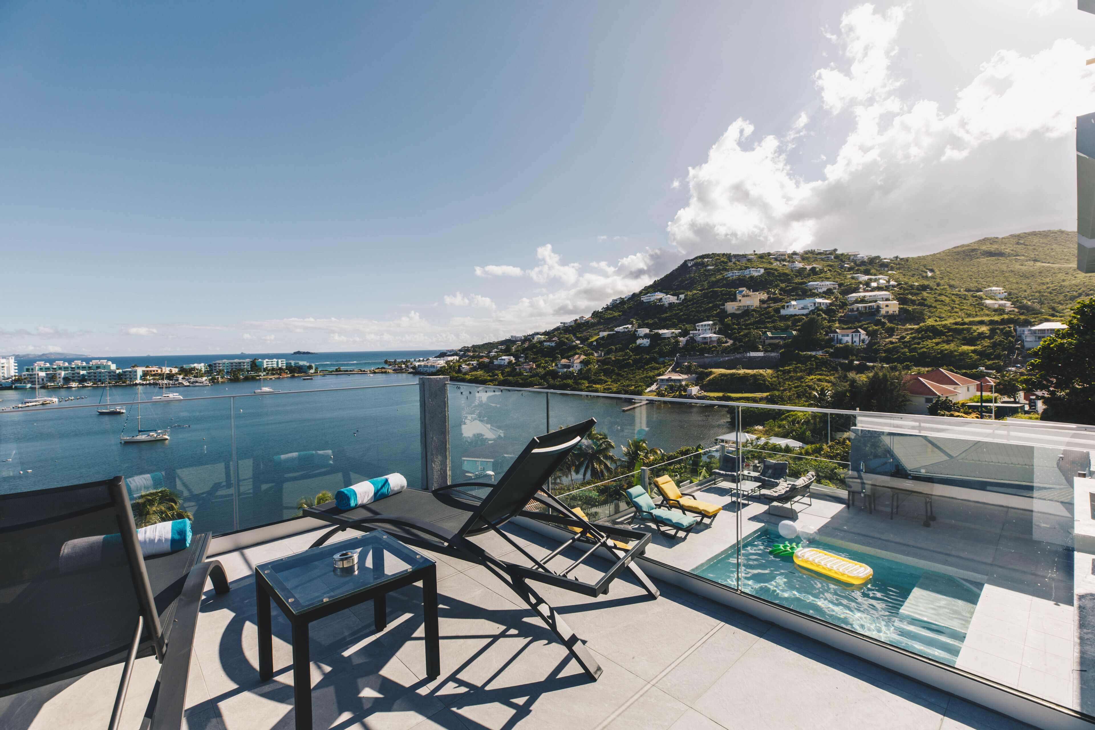 A rooftop pool area with lounge chairs and a glass balustrade overlooking a marina and hillside residences.