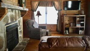 A cozy living room with a stone fireplace, a leather sofa, a patterned armchair, a bookshelf, and a television mounted on the wall.