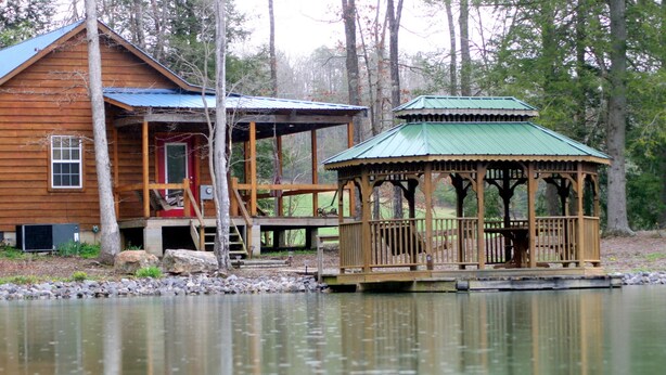 A wooden cabin with a porch and a gazebo by a lake.