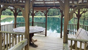 A wooden gazebo with a table and chairs, overlooking a calm lake and surrounded by trees.