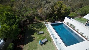 A rectangular swimming pool with a white deck, surrounded by a grassy area and trees. - Seaside Beach House (Marcoola)