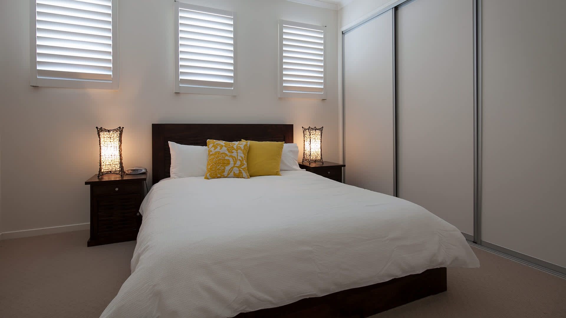 Un dormitorio con una cama grande, dos mesitas de noche con lámparas y tres ventanas con persianas.