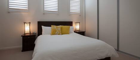 Un dormitorio con una cama grande, dos mesitas de noche con lámparas y tres ventanas con persianas.