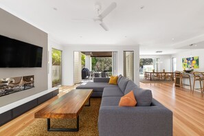 A modern living room with a sectional sofa, a wooden coffee table, a flat-screen TV, and a dining area with a table and chairs. - Seaside Beach House (Marcoola)