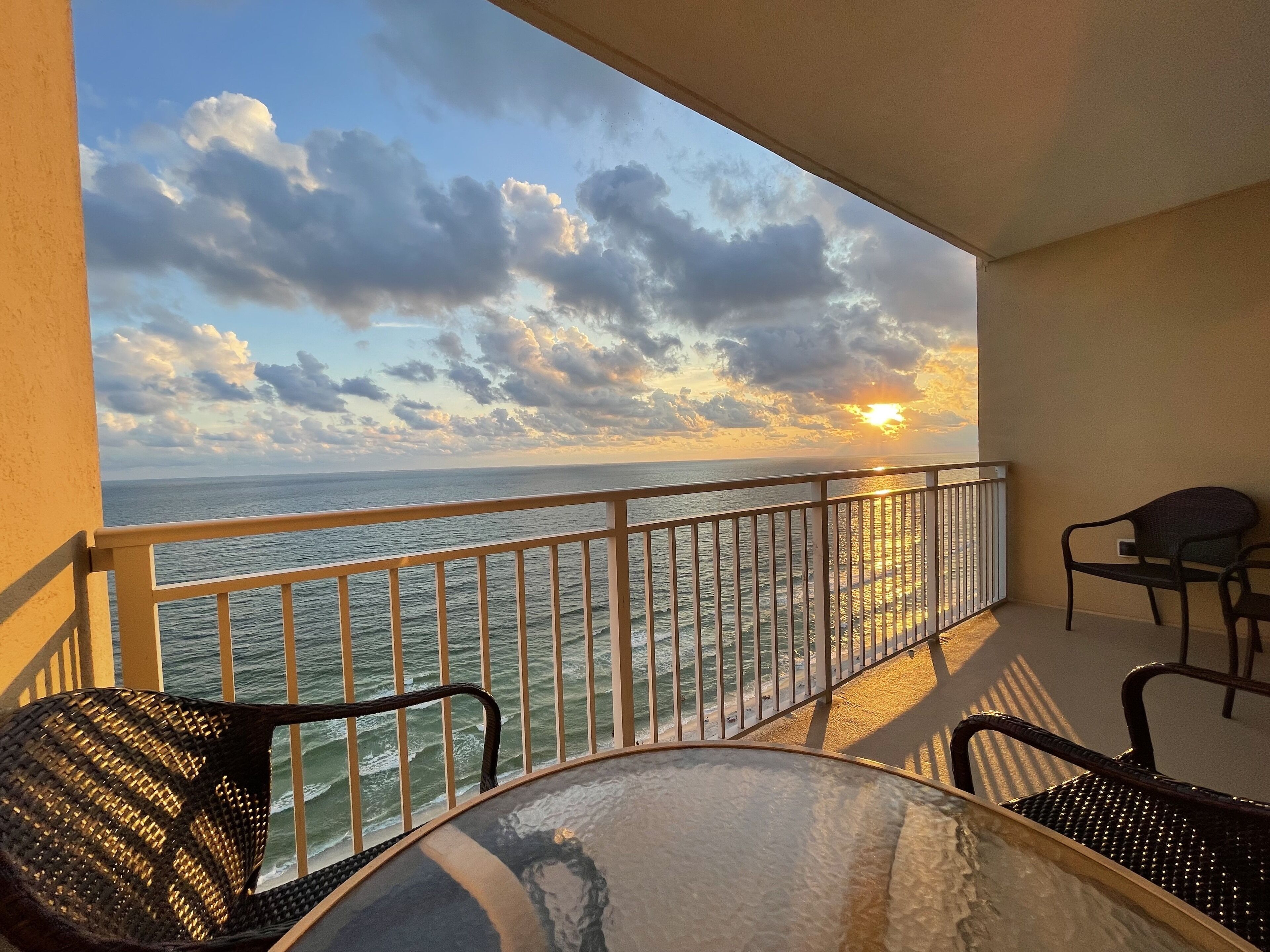 Un balcon avec vue sur la mer et sur le coucher de soleil. Il y a deux chaises et une table.