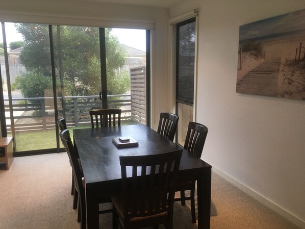A dining area with a large table and chairs, a sliding glass door leading to a backyard, and a painting on the wall. - Coast at The Sands, Torquay (Torquay Surfcoast)