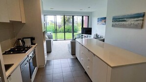 A modern kitchen with a central island, a dining area with a sofa, and a view of the outdoors through large sliding doors. - Coast at The Sands, Torquay (Torquay Surfcoast)