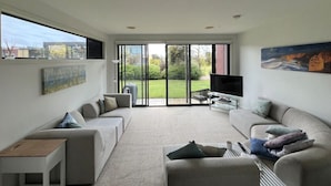 A modern living room with a large sliding glass door leading to a garden, a flat-screen TV, and a sofa set. - Coast at The Sands, Torquay (Torquay Surfcoast)