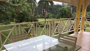 Una mesa con cubierta de vidrio y sillas blancas en un espacio exterior cubierto, con vista a un jardín cuidado.