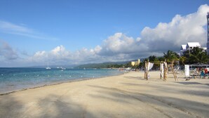 Una playa con cielos despejados, aguas tranquilas y algunas embarcaciones. Se ven palmeras y un edificio al fondo.