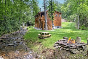 A rustic cabin with a wooden staircase leading to a deck, surrounded by lush greenery and a small stream. - LOG HOME - NEXT TO CREEK / FIREPLACE / HOT TUB / HIKING TRAILS / PET FRIENDLY (Black Mountain)