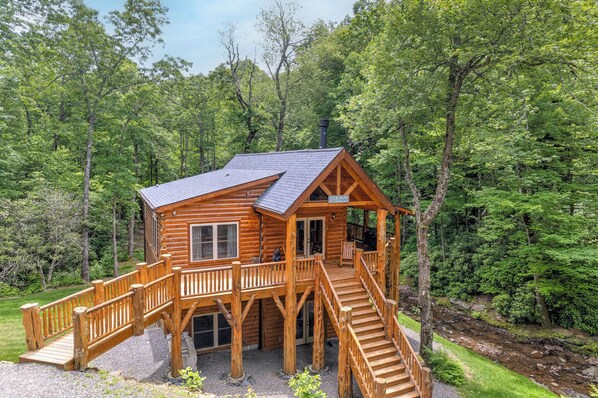 A wooden cabin with a deck and stairs, surrounded by dense forest. - LOG HOME - NEXT TO CREEK / FIREPLACE / HOT TUB / HIKING TRAILS / PET FRIENDLY (Black Mountain)