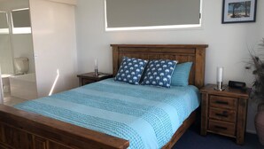 A bedroom with a wooden bed, two nightstands, a bedside table, a window with a beach view, and a potted plant. - 4/42 Manooka Drive - Rainbow Beach. Stunning Panoramic Views and Swimming Pool (Rainbow Beach)