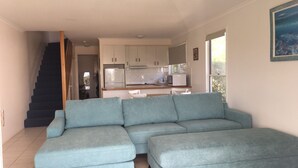 A modern living room with a sectional sofa, a kitchen area with white cabinets and appliances, and a staircase leading to another level. - 4/42 Manooka Drive - Rainbow Beach. Stunning Panoramic Views and Swimming Pool (Rainbow Beach)
