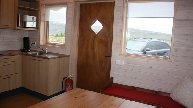 Interior de una cabaña con cocina, una mesa de comedor, un banco rojo y una ventana que ofrece vistas a un coche y montañas.