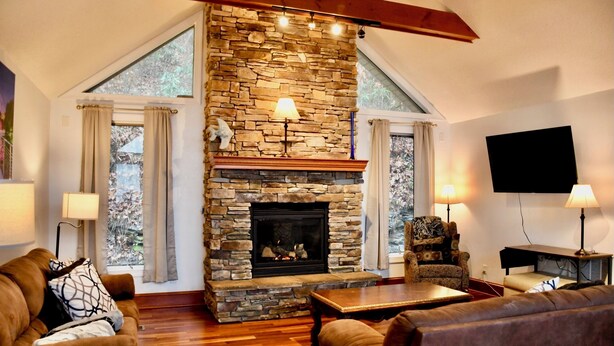 A cozy living room with a stone fireplace, wooden flooring, and a large window.