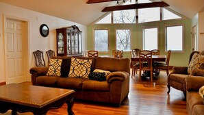 A spacious living room with a high ceiling, wooden floor, a brown sofa set, a dining table with chairs, and large windows.