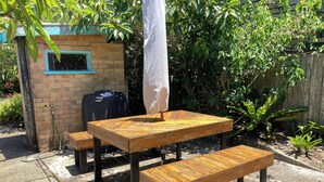Un'area salotto esterna in legno con tavolo e panchina, un tubo da giardino e una piccola costruzione in mattoni con una finestra.