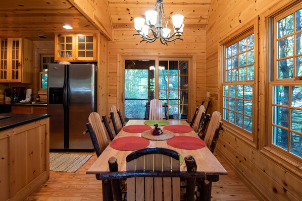 Interior de una cabaña de madera con una mesa de comedor, sillas, un refrigerador y ventanas grandes.