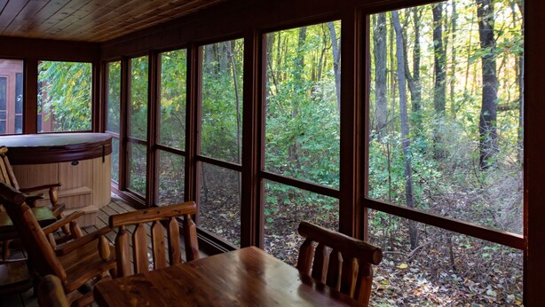 A wooden cabin with a large window overlooking a forest. Inside, there is a wooden table and chairs, and a hot tub is visible through the window.