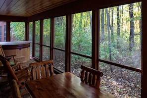 Een houten hut met een groot raam dat uitkijkt op een bos. Binnen staat een houten tafel met stoelen, en er is een hottub zichtbaar door het raam.