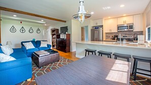 A modern living room with a blue sofa set, a wooden coffee table, and a dining area with a large table and bar stools. - Perfect Location, Historic Town Ctr: Ground 4 Bed 3 Bath Condo (Provincetown)