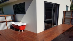 Een modern terras buiten, met een rood bad, houten relingen en een glazen deur die leidt naar een overdekt gedeelte.