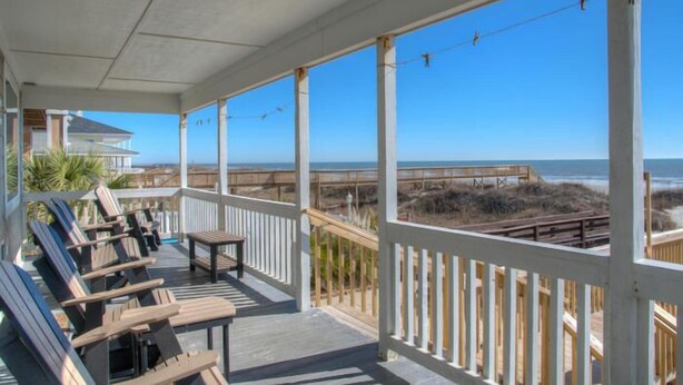 A covered outdoor area with wooden deck chairs and a table, overlooking a beach and boardwalk.