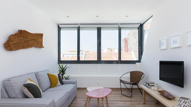 A modern living room with a grey sofa, a small round table, a wooden chair, a TV, and a large window with a city view. - A pearl of an apartment in Belém (Lisboa)