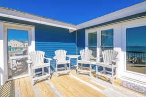 Um terraço com quatro cadeiras Adirondack brancas e uma mesa pequena, com vista para um edifício azul e o oceano.