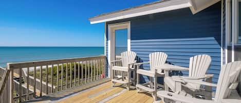 A wooden deck with white chairs and a table overlooking a beach.